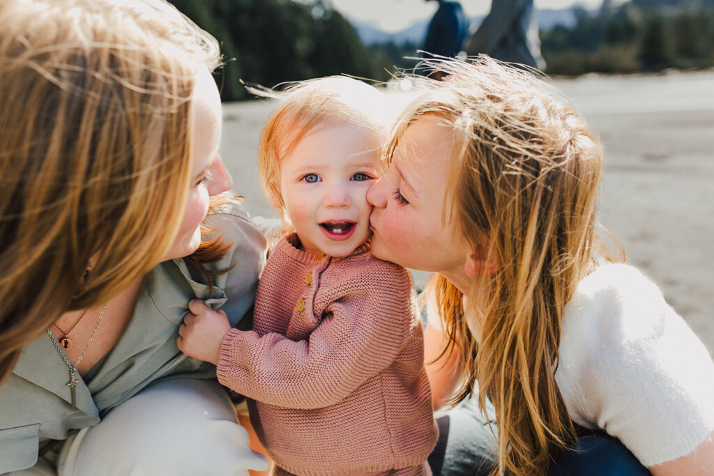 three girls on the beach on a windy day. The middle sister is kissing her toddler sister on the cheek while the oldest sister looks on