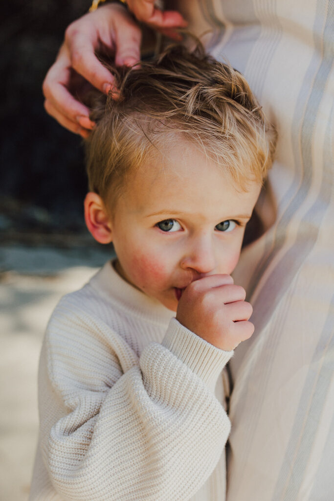 A toddler child snuggled into his mother's side. He is sucking his thumb and looking into the camera mischeviously