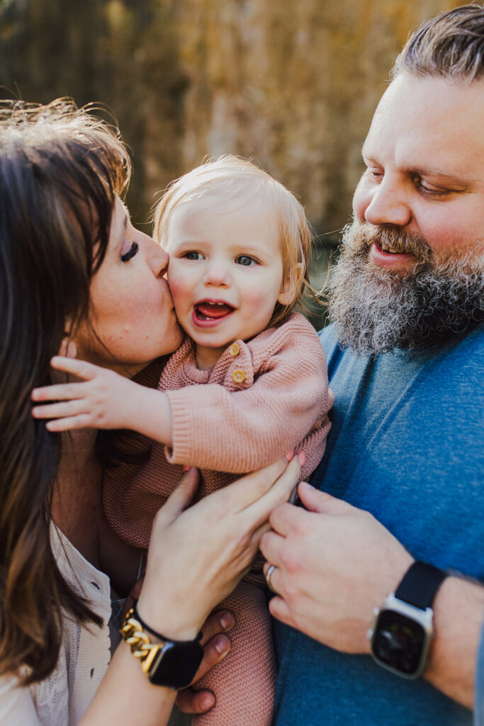 a picture of a mother and father and their toddler daughter. The mother is kissing her daughter on the cheek