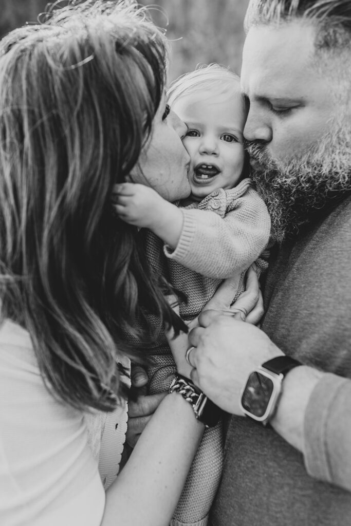 A mother and father holding their toddler daughter between them. They are both kissing her on the cheek