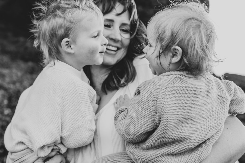 A black and white image of mother holding her two young children in her arms. They are smiling at each other. 
