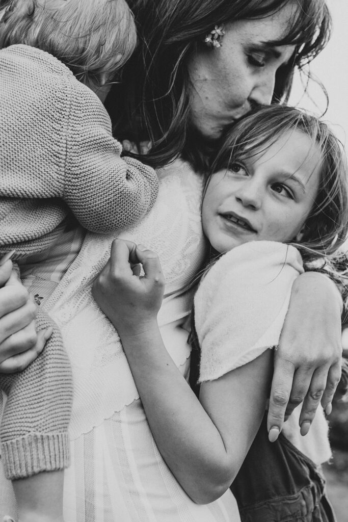 A mother holding a toddler with an arm around her older daughter. The mother is kissing the top of her older daughter's head