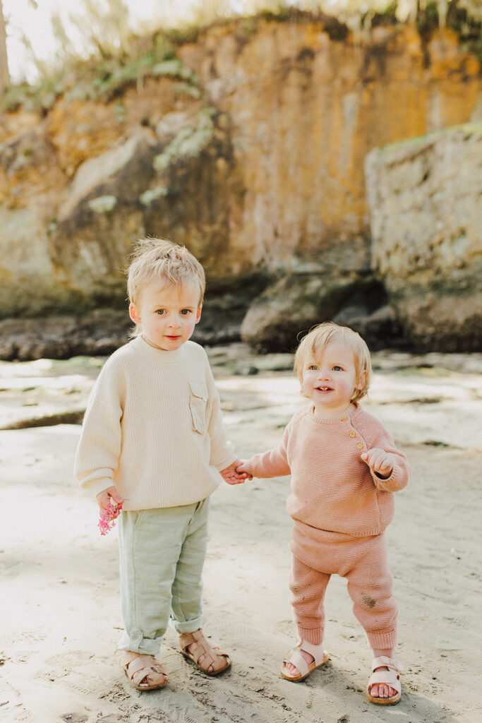 A toddler boy and girl holding hands on the beach with the sea stack at Salt Creek beach behind them. 