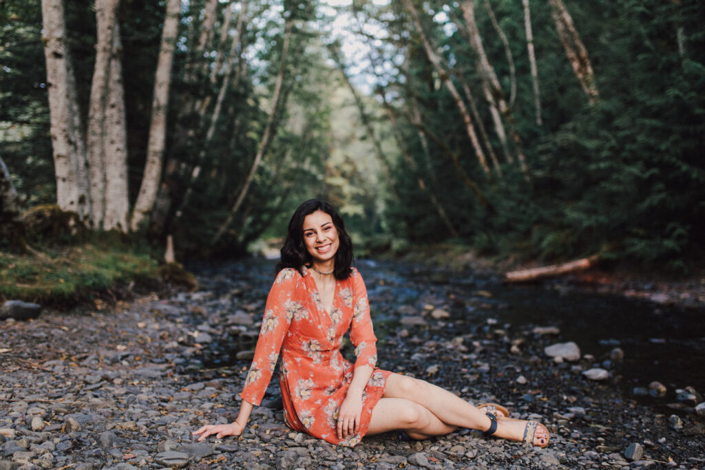 A teenage girl sitting by a creek in the woods. She's wearing an orange dress.