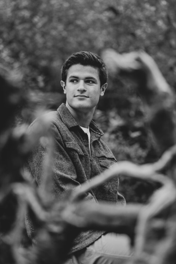 A black and white image of a teenage boy sitting on a driftwood log. 