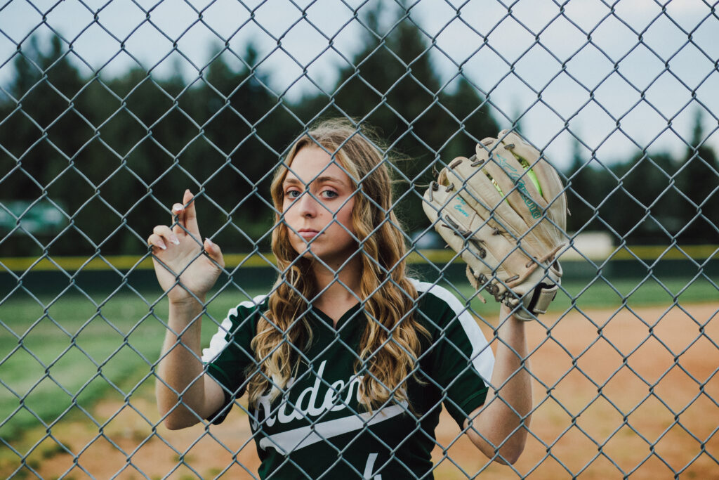 A girl looking through a chan link fence at a softball field