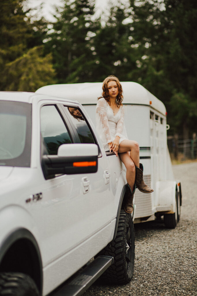A girl sitting on the back of her truck that is hitched up to her horse trailer.