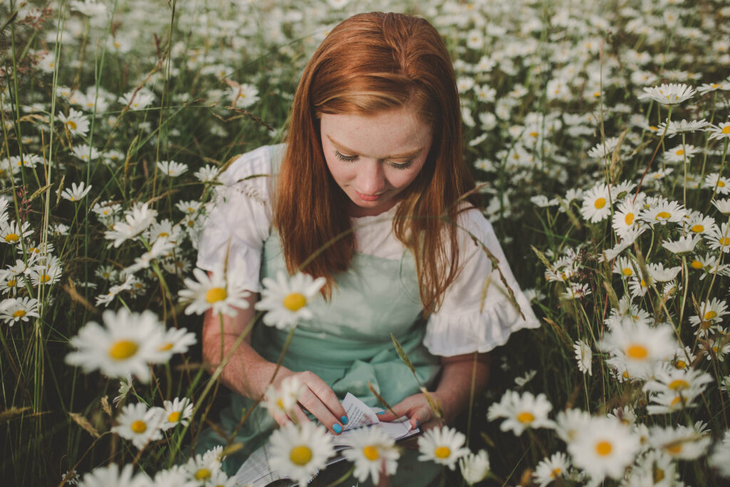 A teenage girl sitting in a daisy field at sunset while reading a book.